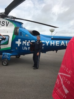Man standing beside a blue Mercy Health helicopter on a runway, with a red cloth in the foreground