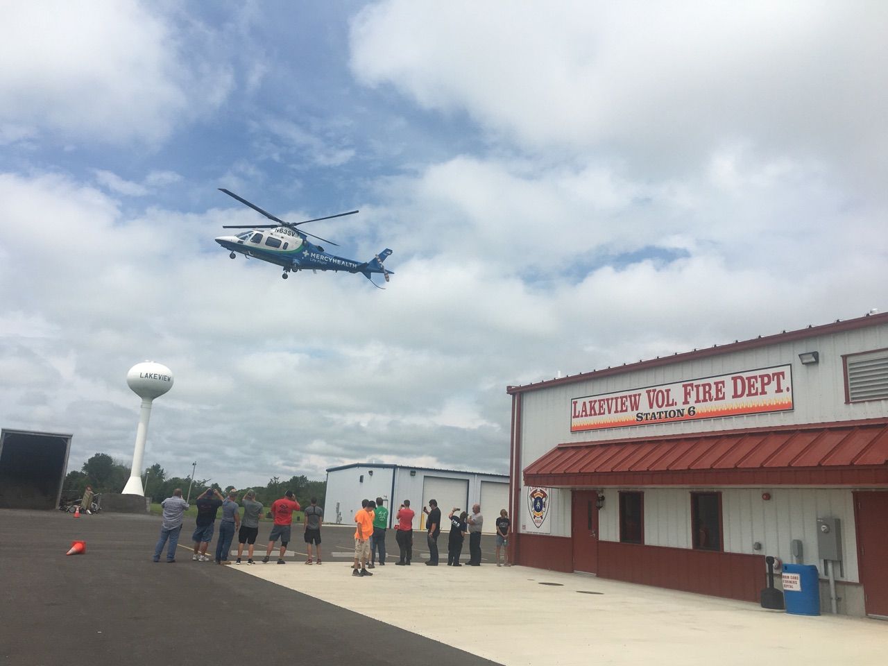 Helicopter hovering above a fire station with firefighters gathered outside under cloudy skies