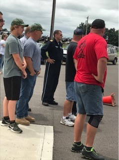 Group of people standing outdoors in a parking lot, listening to a man in a black tracksuit beside traffic cones.