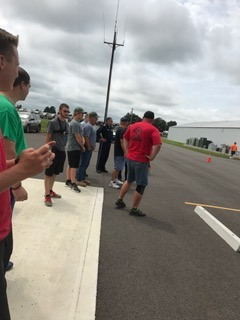 People standing along a roadside near a parking lot, watching traffic cones under a cloudy sky.