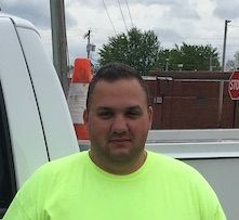 Man in neon yellow shirt standing outdoors near a white van and brick wall under cloudy sky