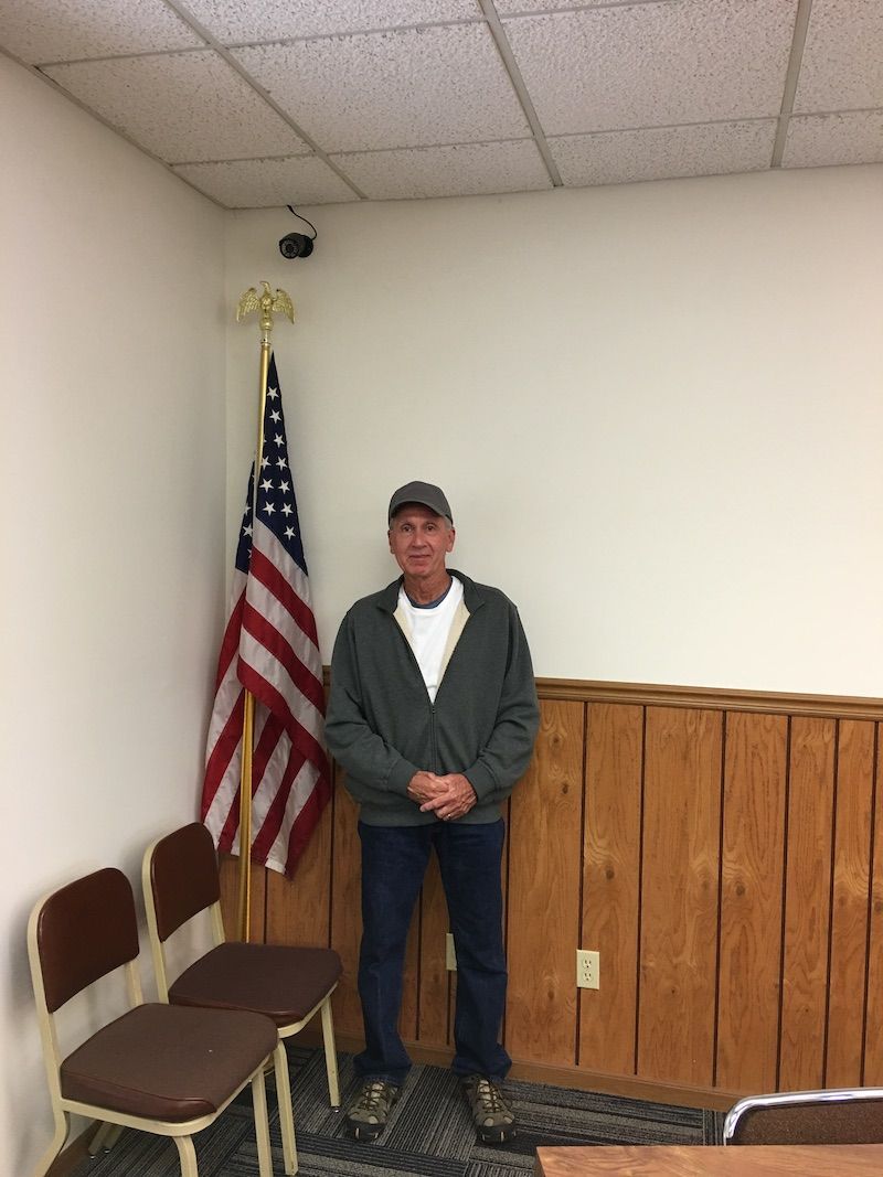Man standing beside an American flag in a room with chairs and wood paneling