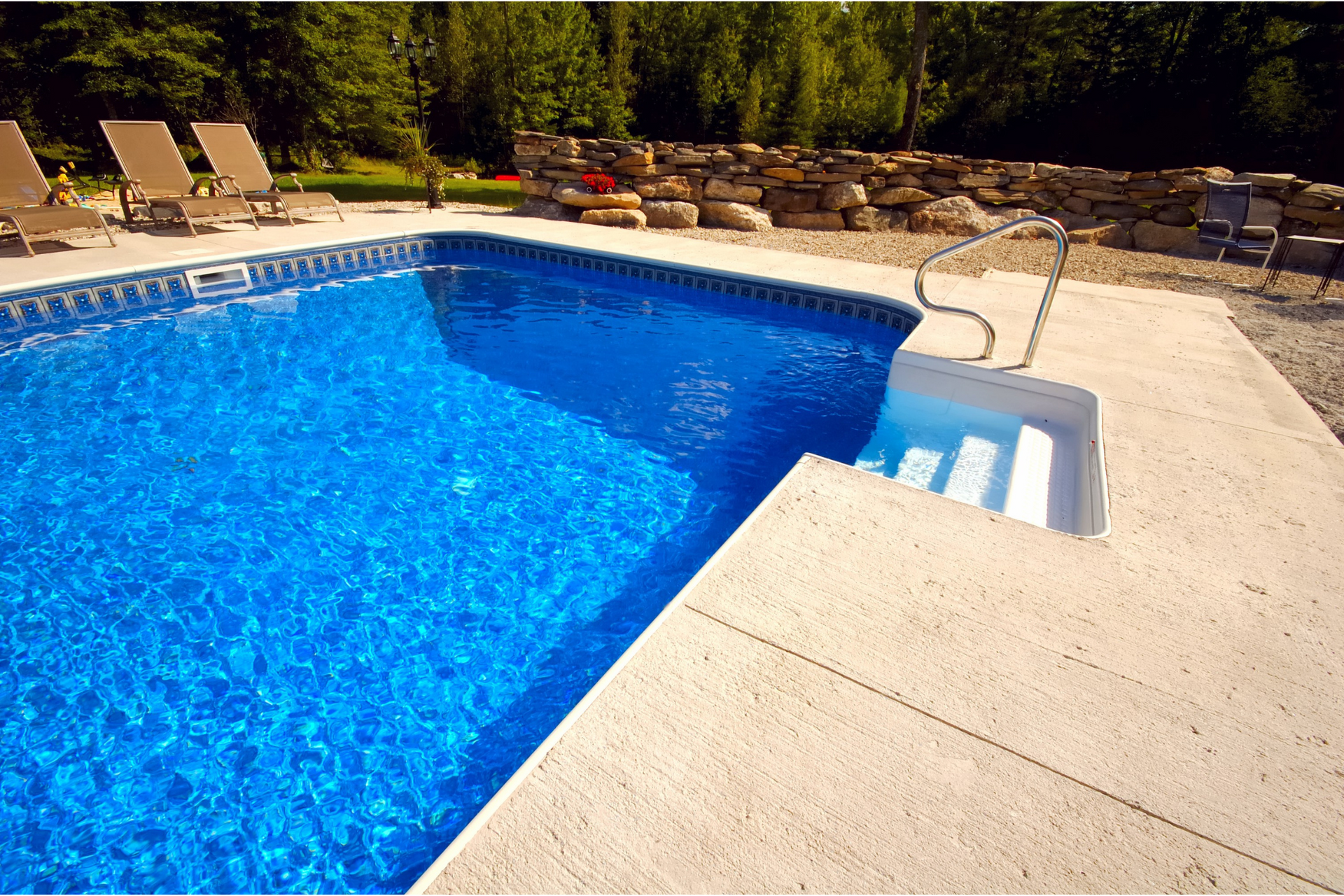 Blue rectangular swimming pool with steps, surrounded by concrete and trees.