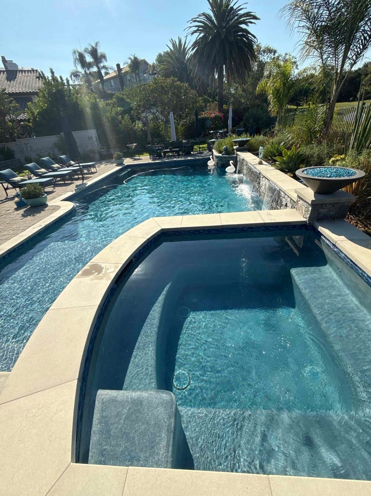 Pool and spa with blue water, surrounded by tan stone and lush landscaping. Sunny outdoor setting.