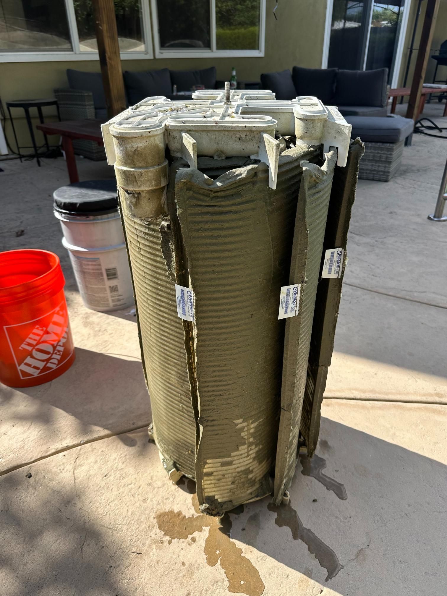 A dirty swimming pool filter on a patio next to a red bucket and clear container.