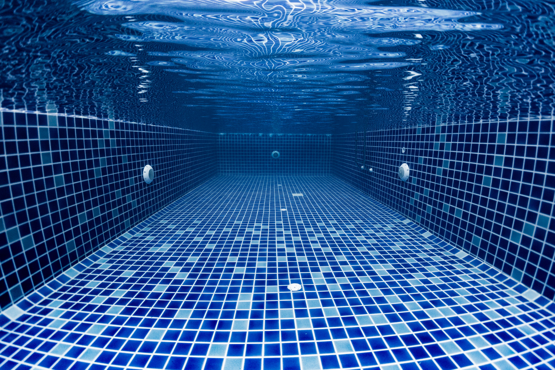 Underwater view of a tiled, empty swimming pool. Dark blue tiles create a mosaic pattern.