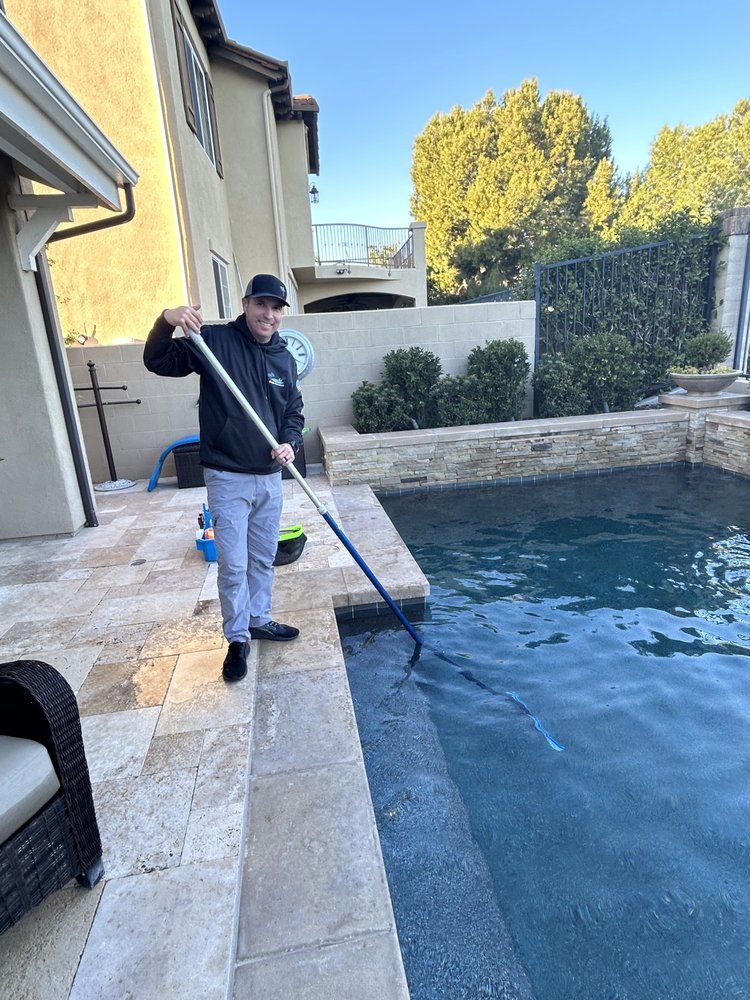 Man cleaning a pool with a net, wearing a black jacket and hat, standing on a patio.