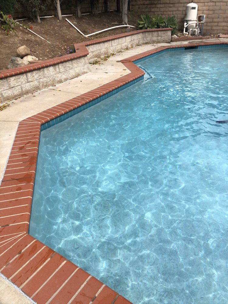 A blue swimming pool with brick edging. The pool is surrounded by a concrete patio and a retaining wall.