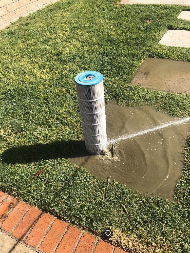A cylindrical pool filter spewing water into a muddy patch on a lawn. Brick border in the foreground.