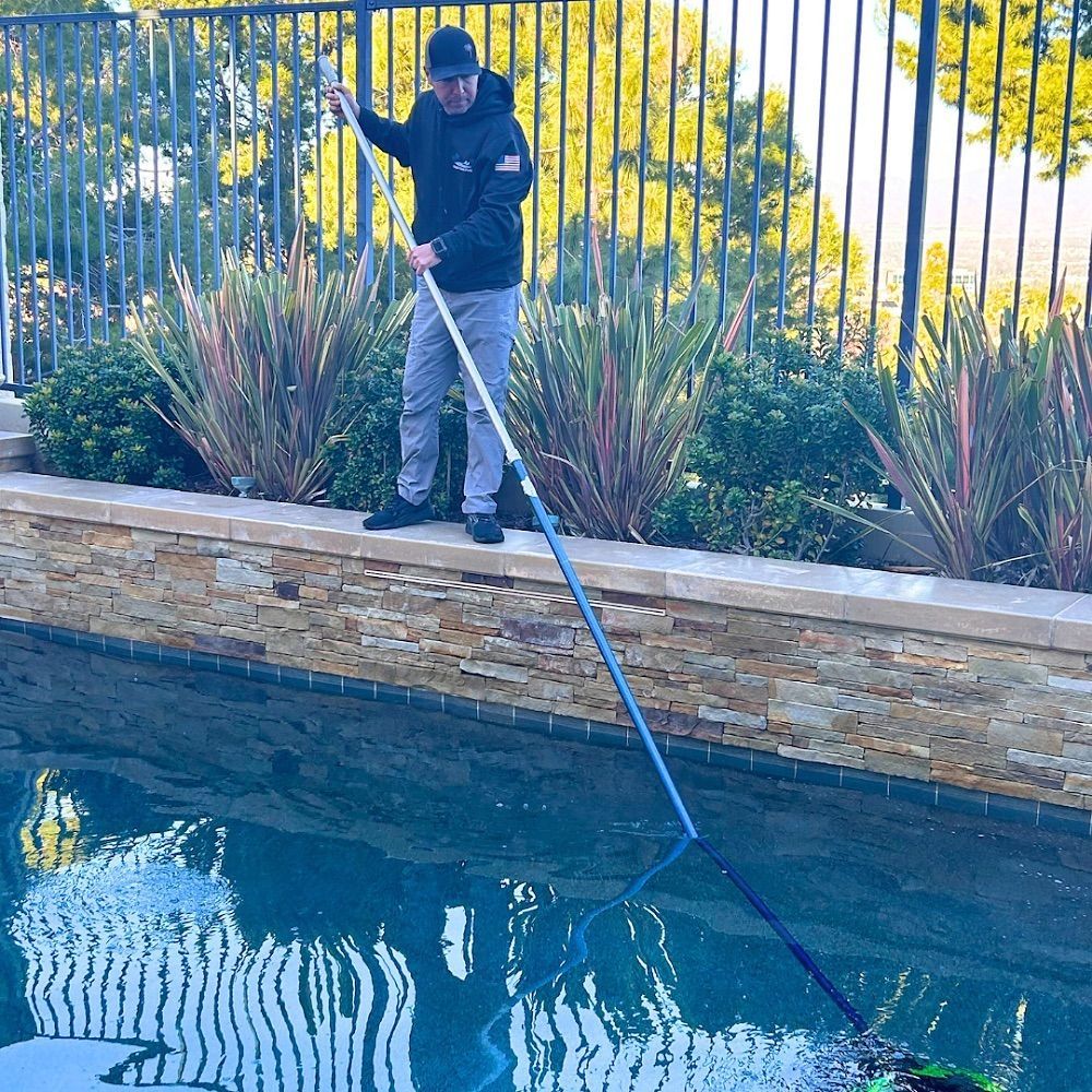Man in black jacket cleans a pool with a net, standing near a fence and plants.