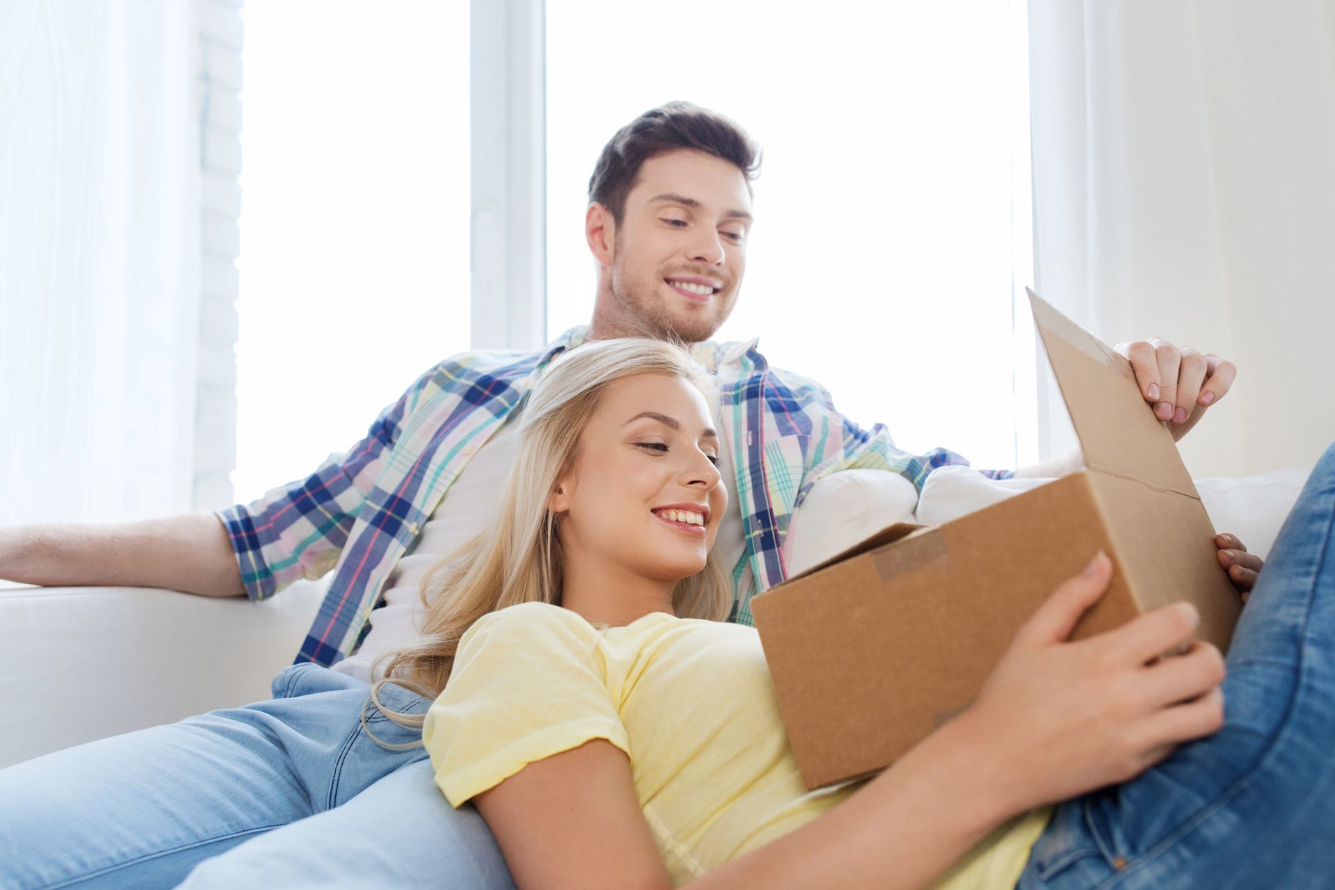 Pareja en un sofá abriendo una caja de cartón, sonriendo. Habitación luminosa con luz natural.