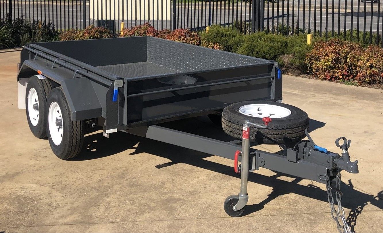 Gray tandem-axle trailer with white-rimmed tires, spare tire, and open bed, parked outdoors.