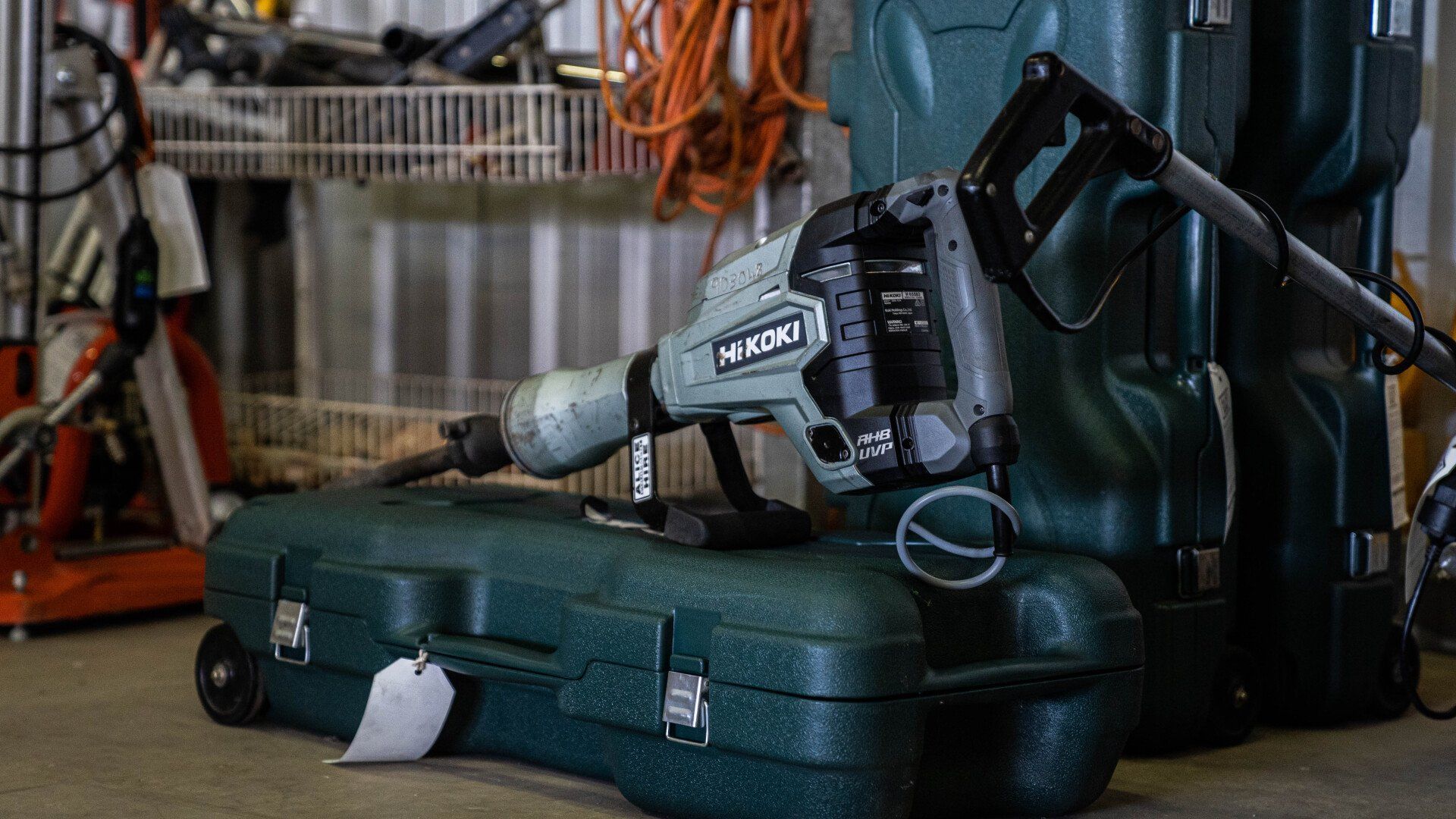 A jackhammer on a green case in a cluttered storage area with tools and shelving.