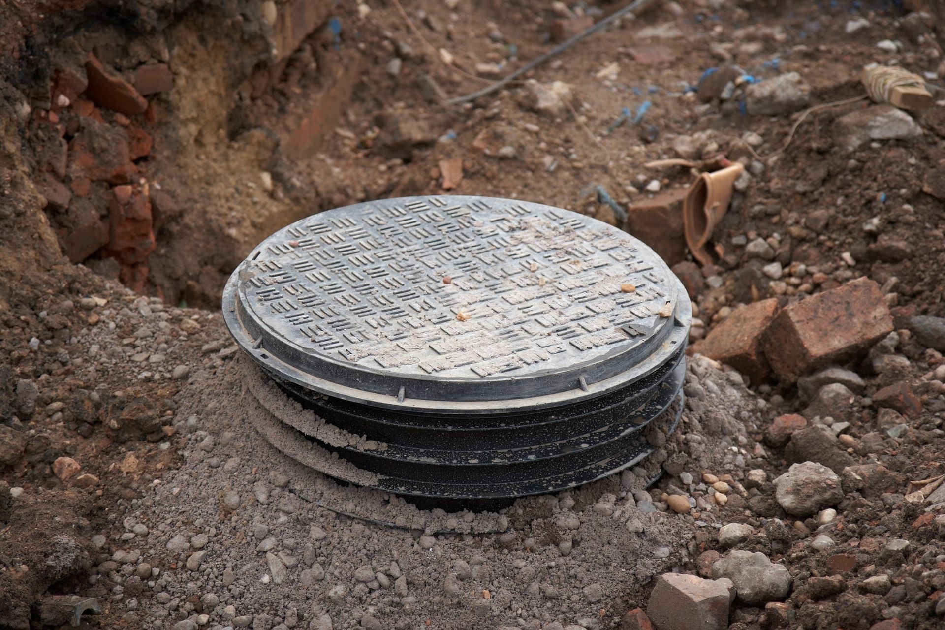 A round, black manhole cover sits atop a cylindrical structure surrounded by dirt, bricks, and debris.