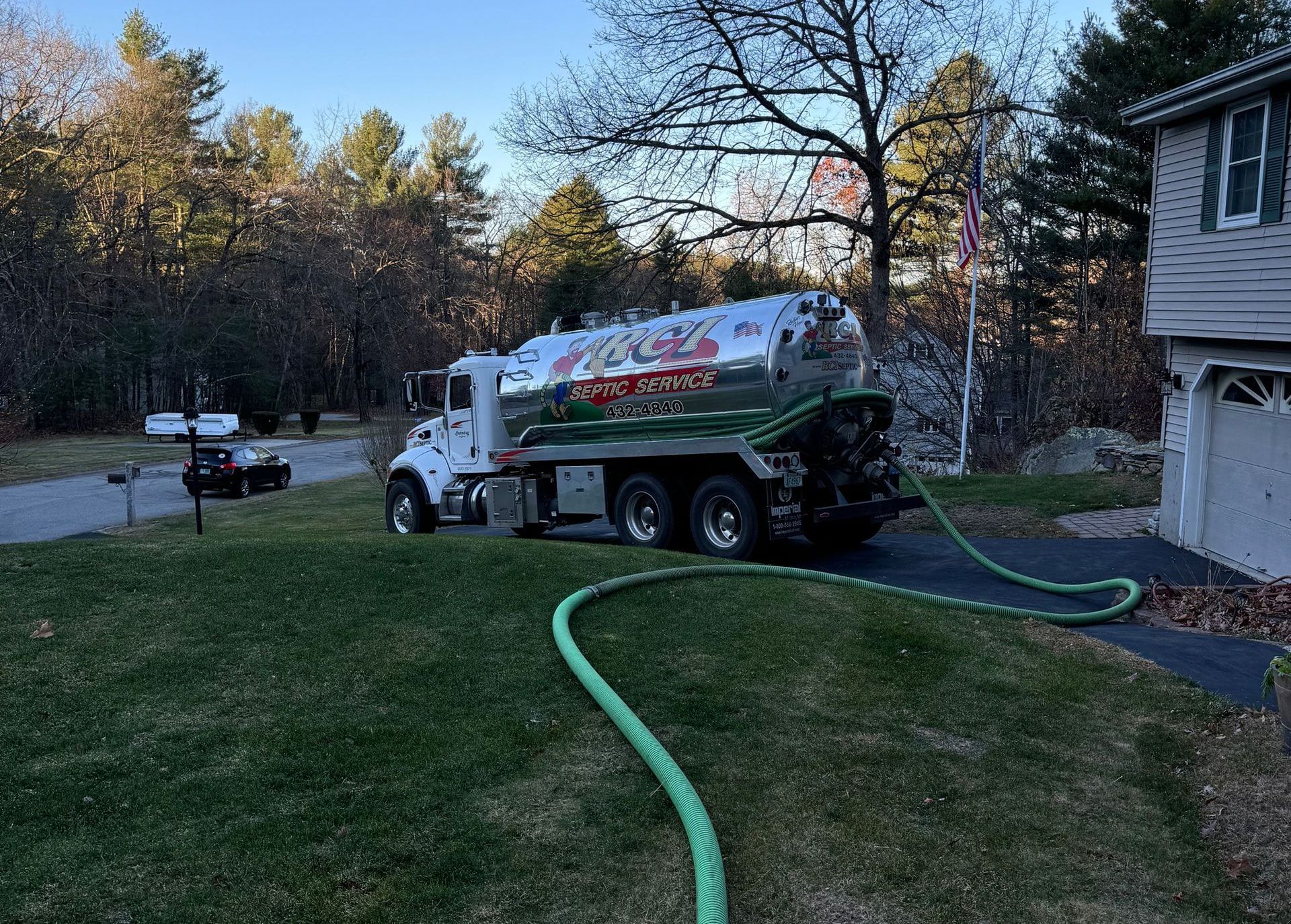 A septic tank truck parked on a residential lawn with a hose extending towards a house; green grass and trees in the background.
