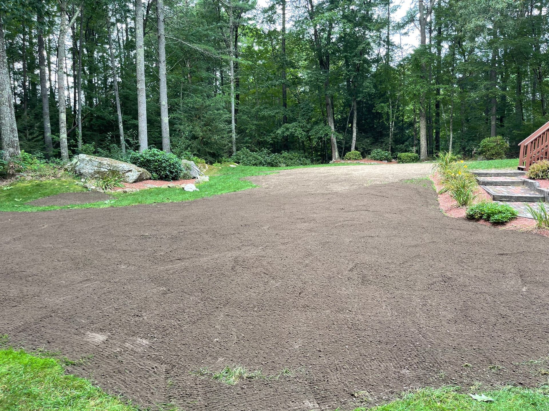 A cleared yard area, covered in dark soil, ready for planting with a forest backdrop and steps to a house.