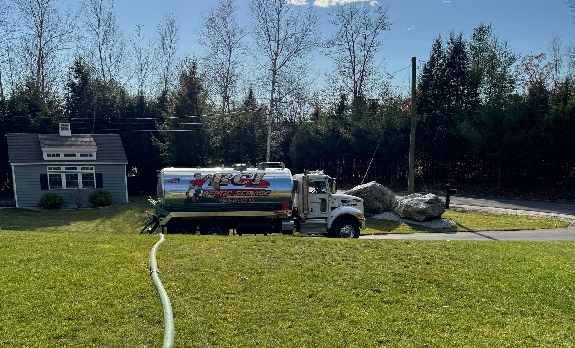A septic tank truck parked on a grassy lawn next to a small house. A hose stretches toward the house.