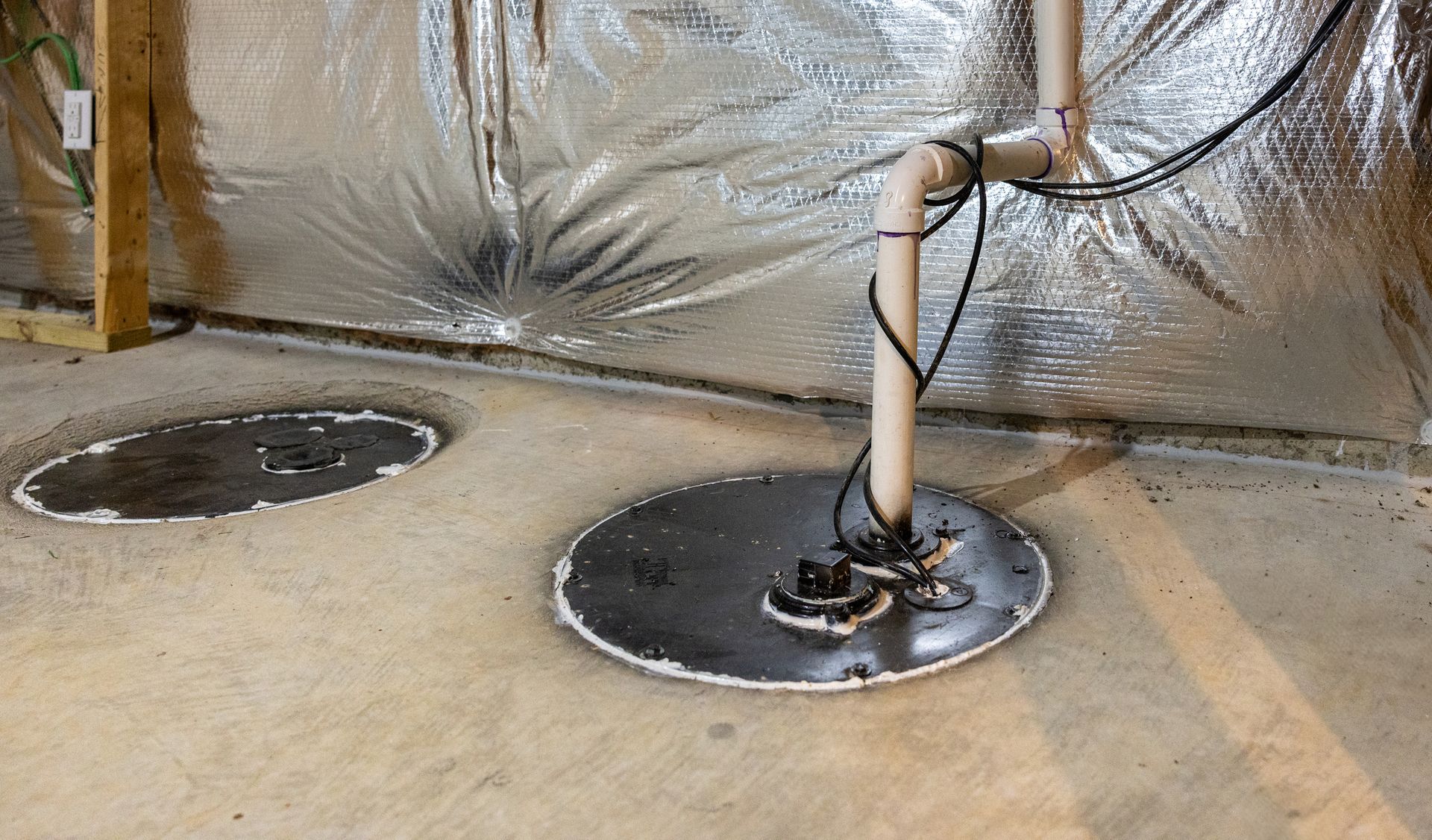 Basement floor with two open sump pit covers and a vertical pipe against a reflective insulated wall