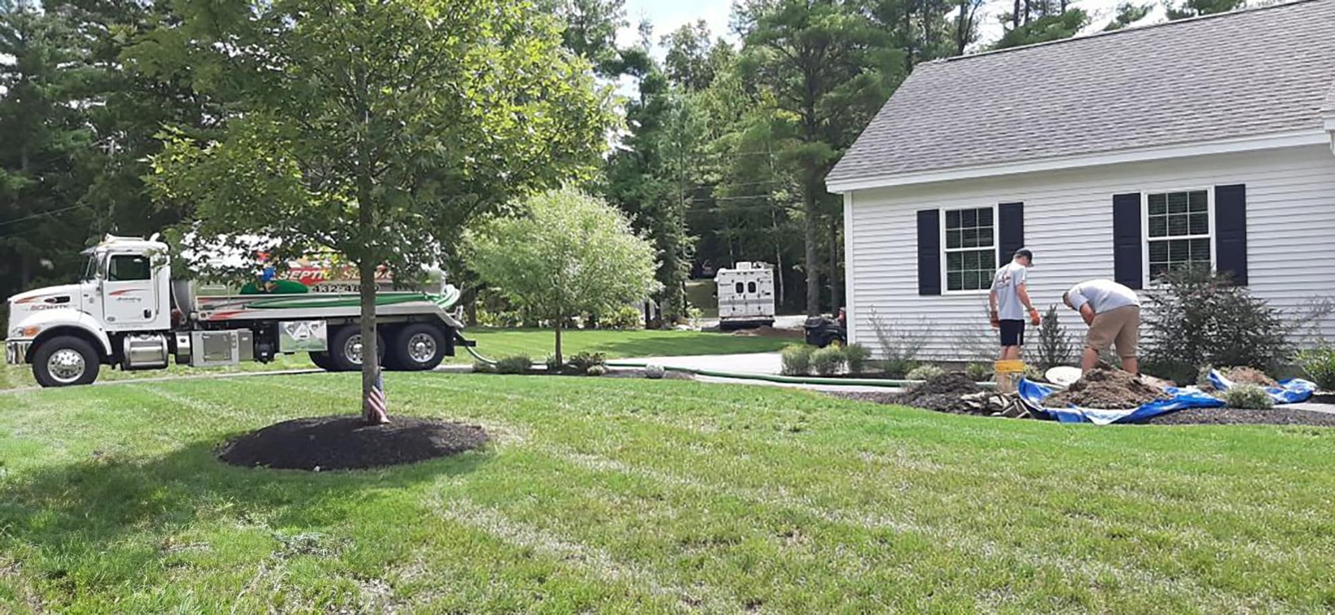 A white truck and two men working on landscaping in front of a small house with blue shutters. The yard is green with a tree.