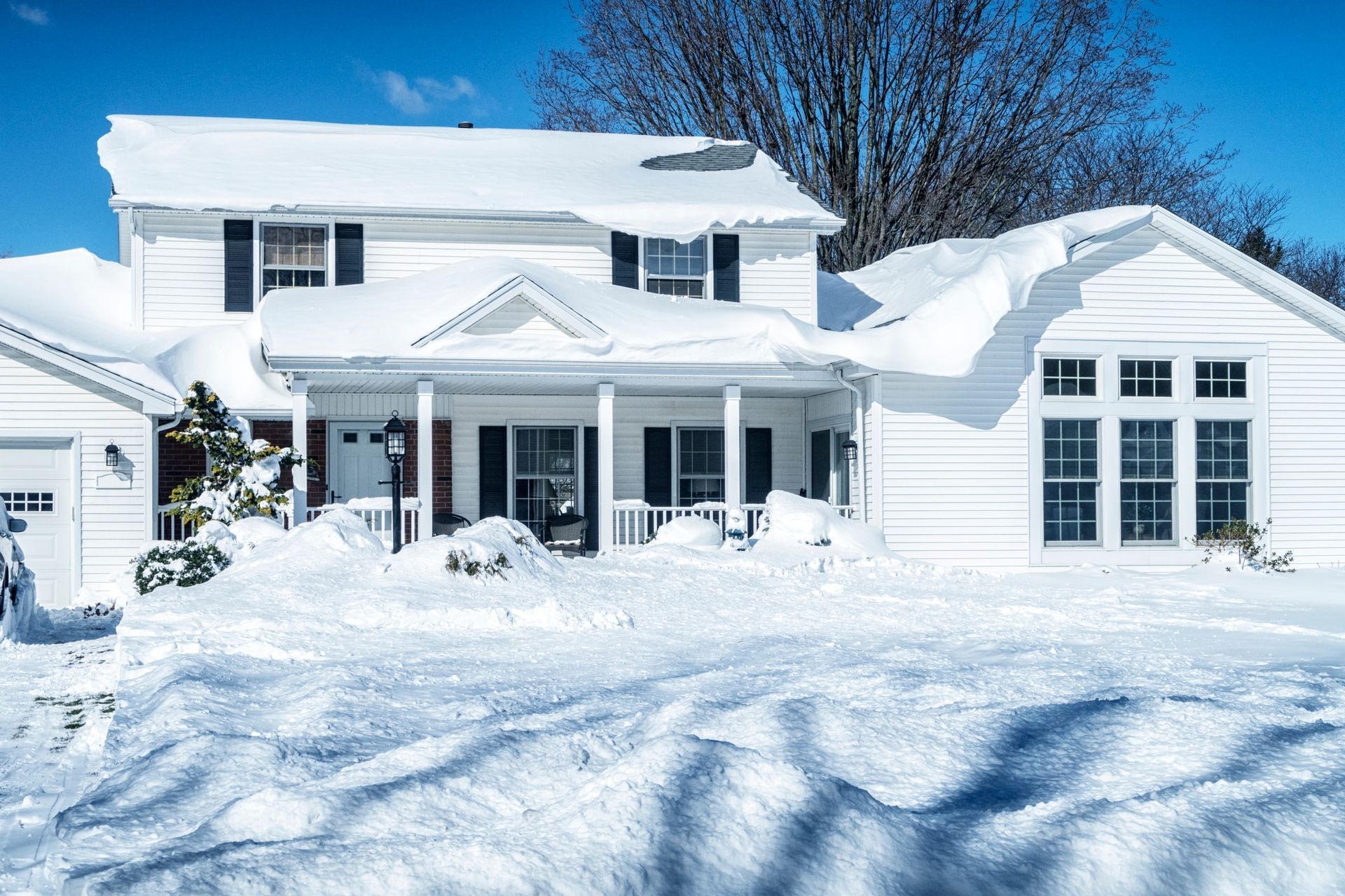 White house covered in deep snow, with porch and garage visible on a sunny winter day.