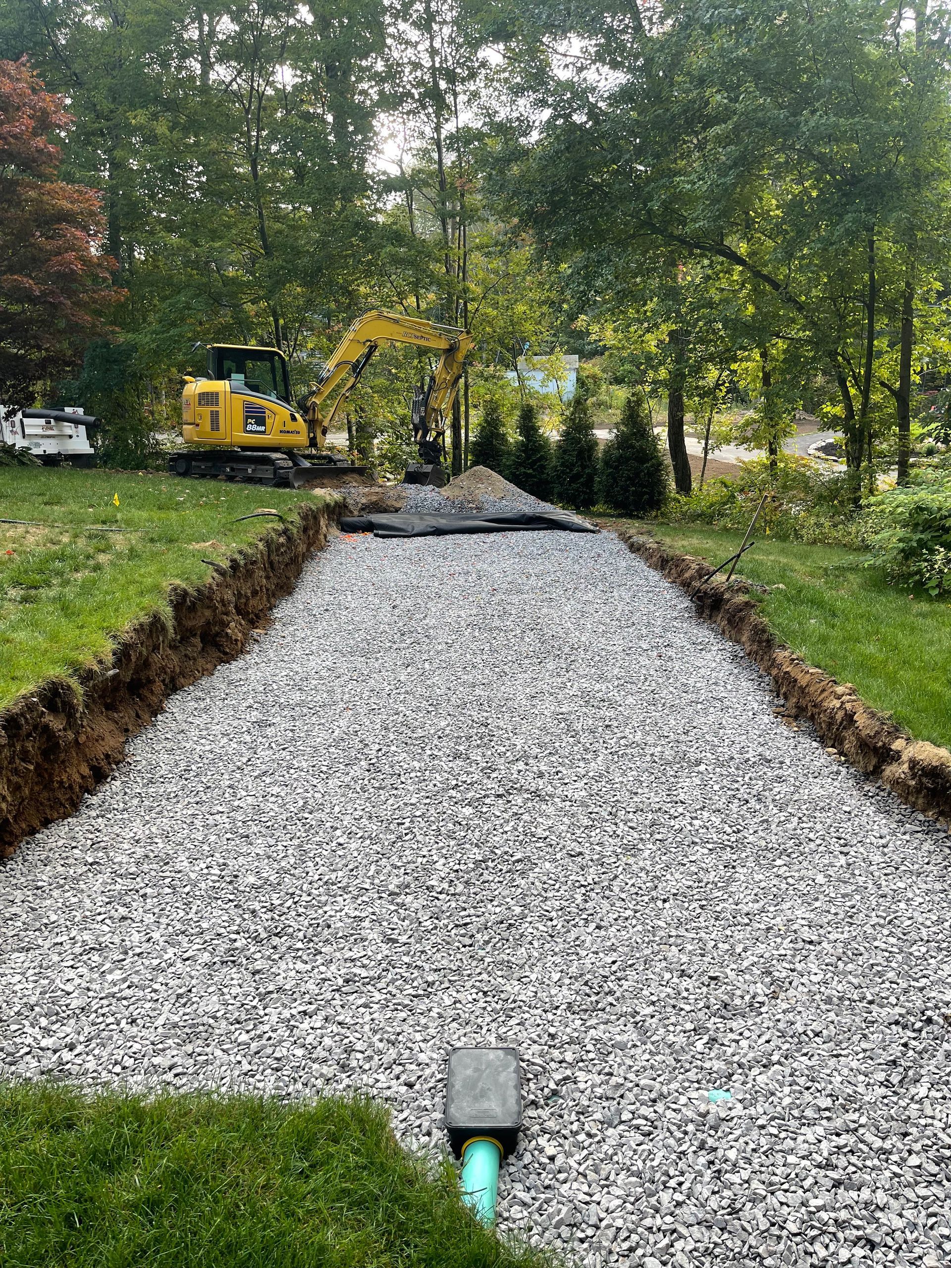 Gravel-filled drainage trench under construction beside a grassy yard, with a yellow excavator in the background