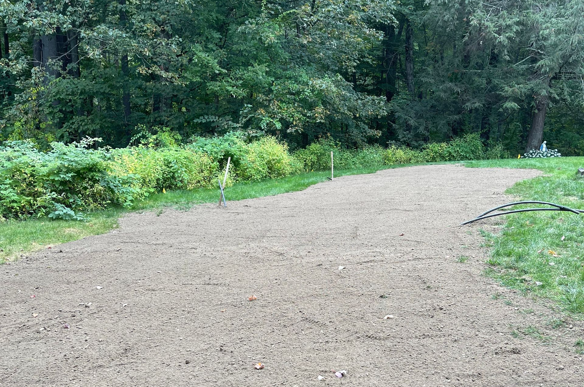 A freshly tilled, gravelly area for planting sits between green grass and a border of wild plants, with trees in the background.