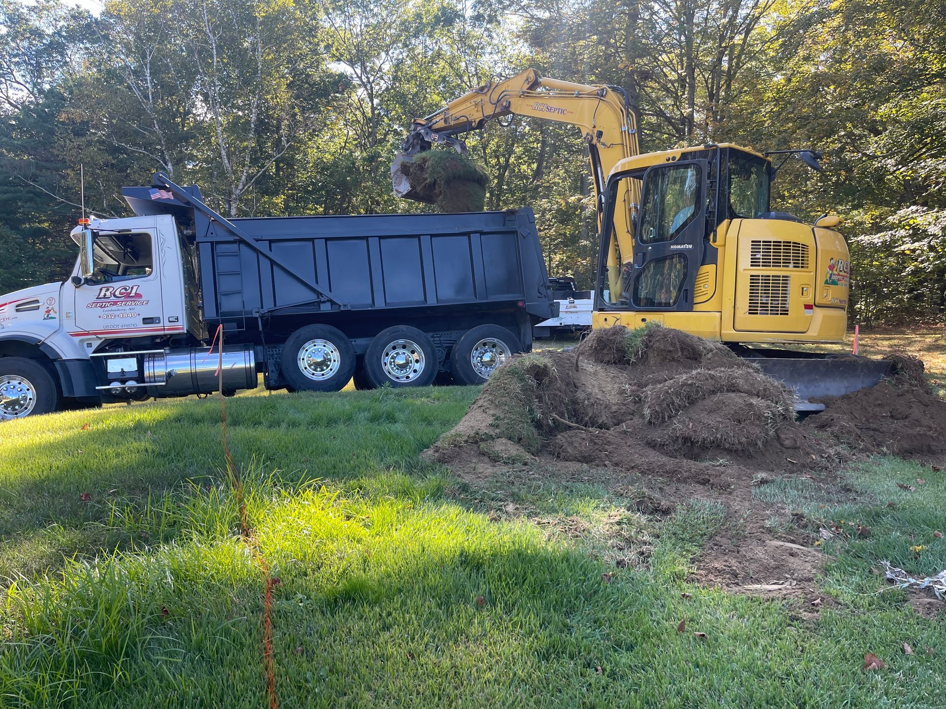 Yellow excavator loading a black dump truck with dirt in a grassy field.
