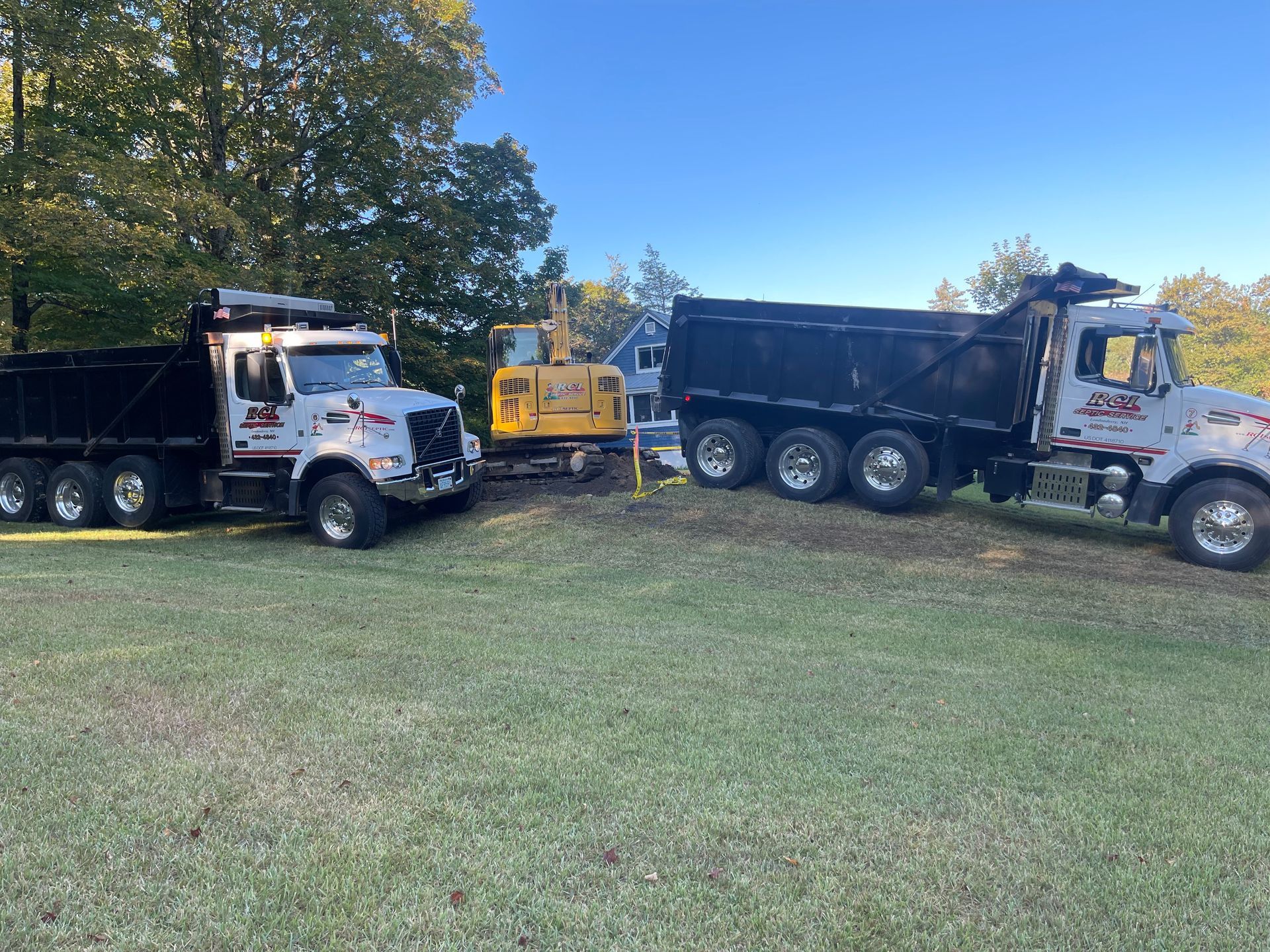 Two white dump trucks and a yellow machine parked on grass under trees