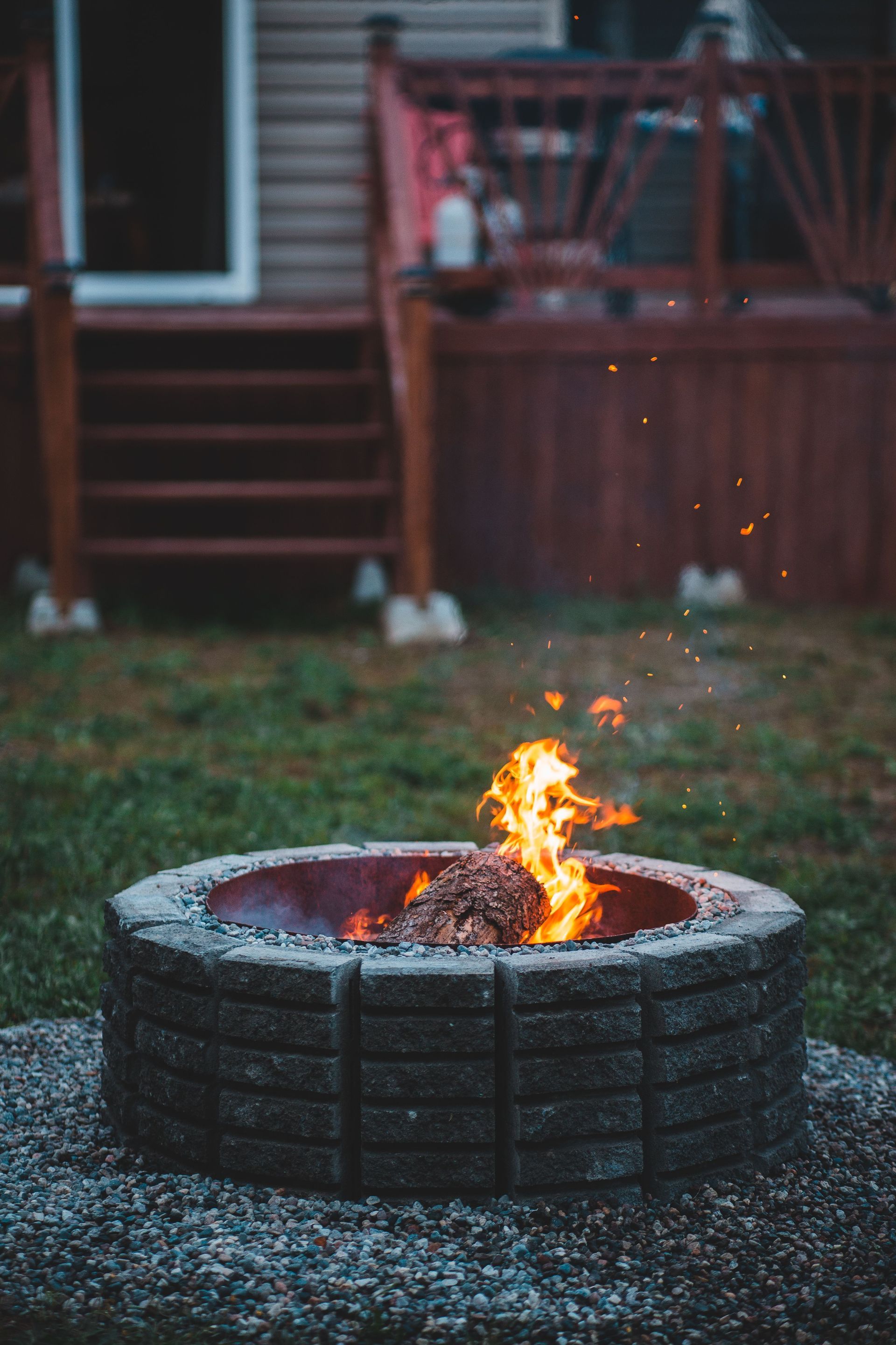 Backyard fire pit with bright flames on gravel in front of a wooden deck and stairs