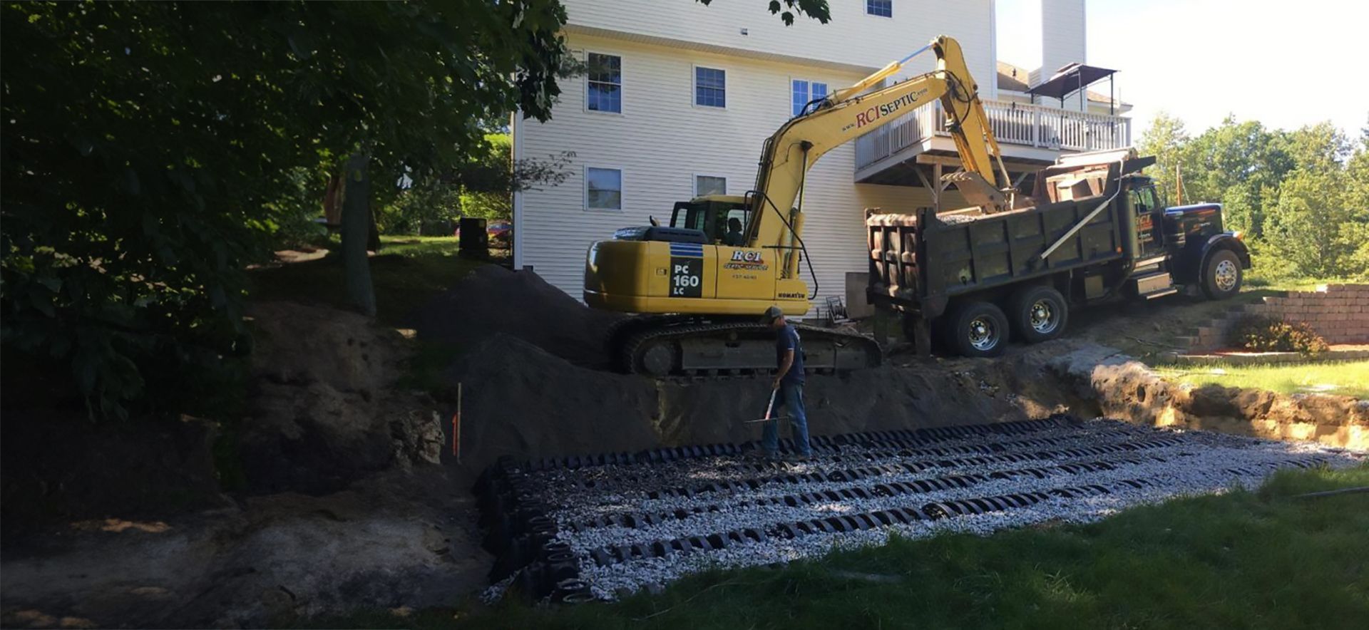 a yellow excavator is loading dirt into a dump truck in front of a house that is getting a septic system installed