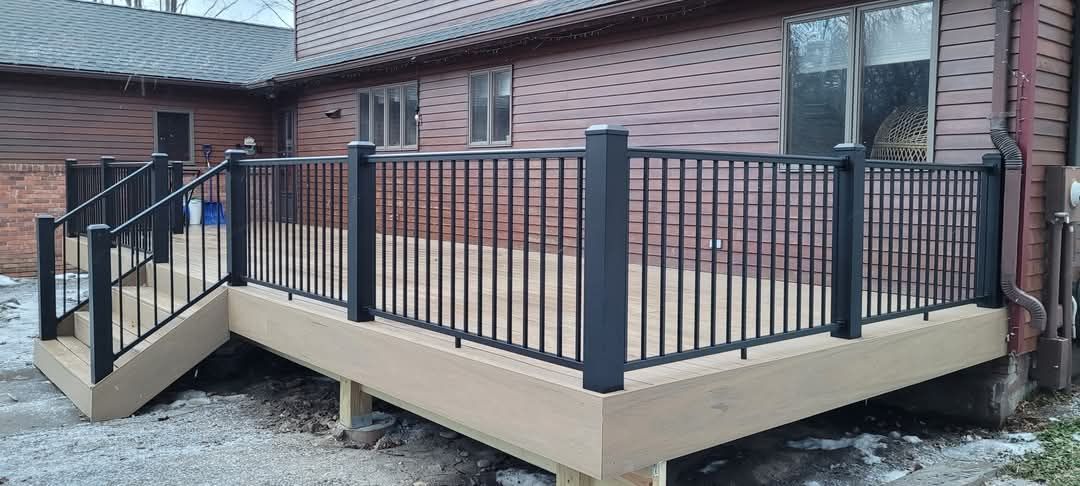 A wooden deck with a metal railing is in front of a brick house.
