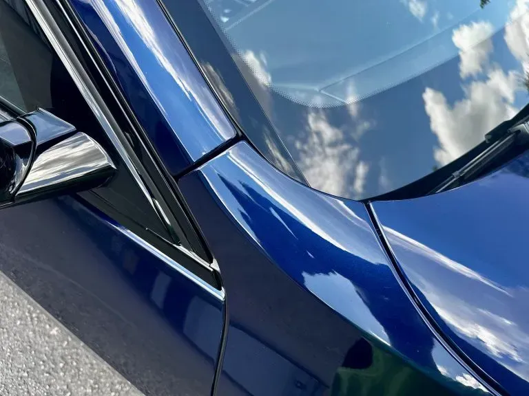 Close-up of a blue car’s side mirror, door seam, and windshield reflecting clouds.