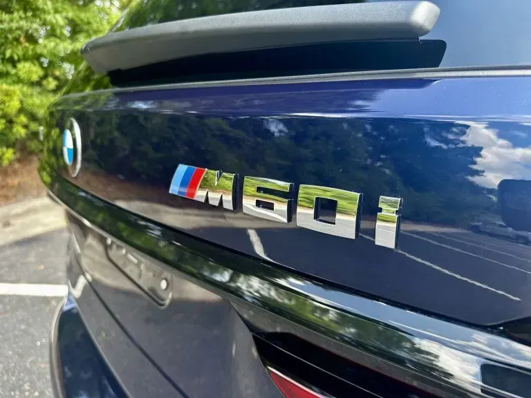 Close-up of a blue BMW M50i badge on a car trunk with trees reflected in the paint