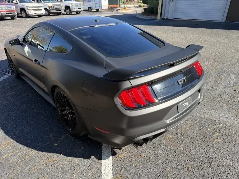 Matte black Ford Mustang coupe parked in a lot, viewed from the rear-left side.