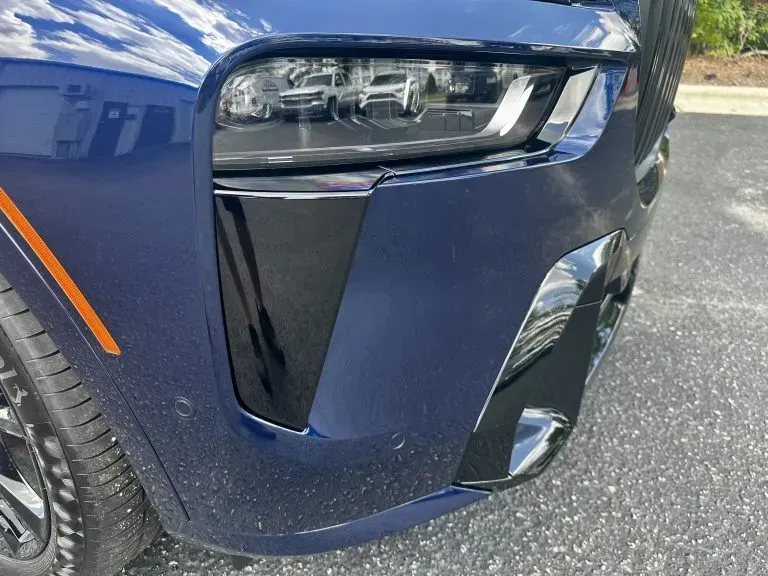 Close-up of a blue car’s front corner, showing the headlight, bumper, and wheel on pavement.