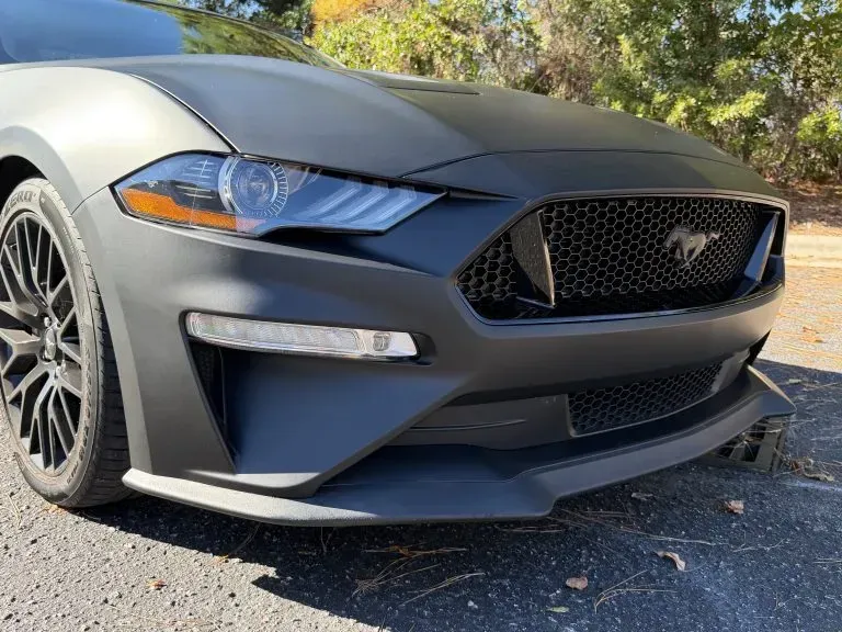 Front view of a gray Ford Mustang parked on a sunny street, with trees in the background