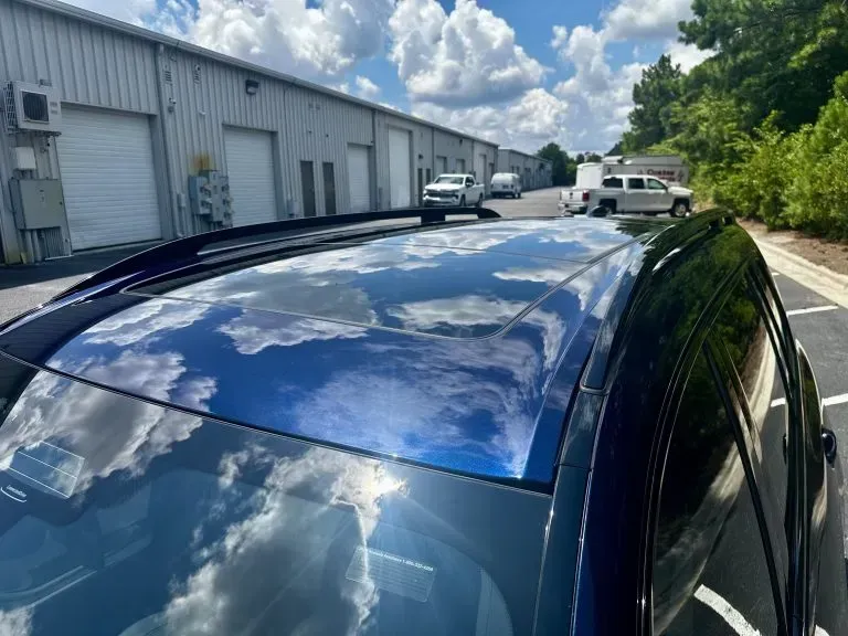 Blue car with sunroof reflecting clouds, parked beside industrial buildings under a sunny sky.