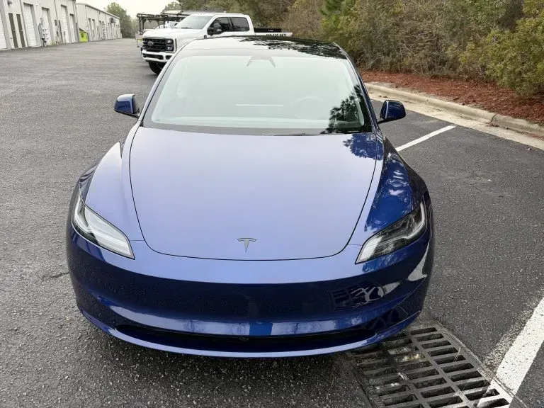 Blue Tesla sedan parked in a lot, front view with another car behind it.