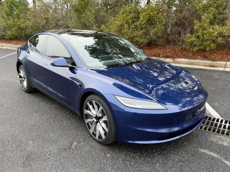 Blue sedan parked on a street beside a curb and storm drain, with trees in the background