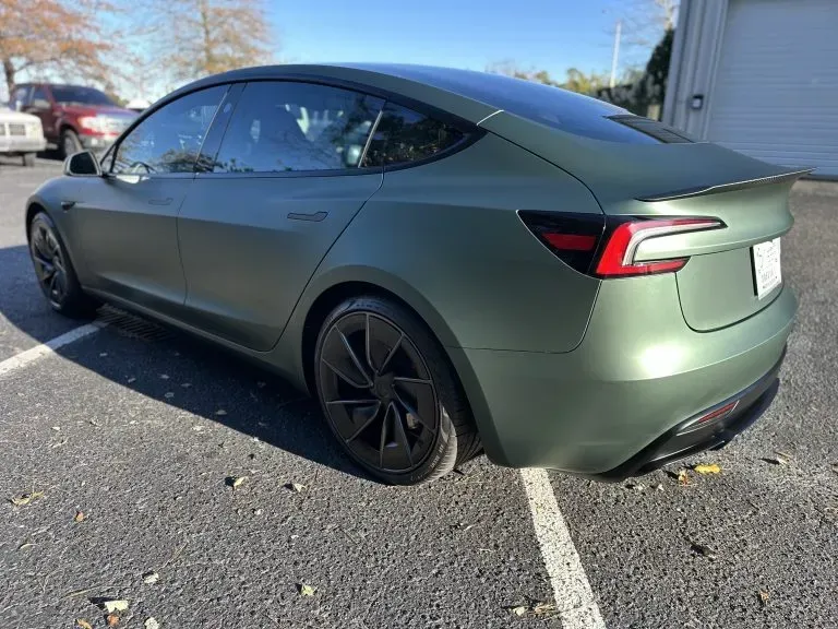 Green Tesla sedan parked in a lot, rear side view on a sunny day.