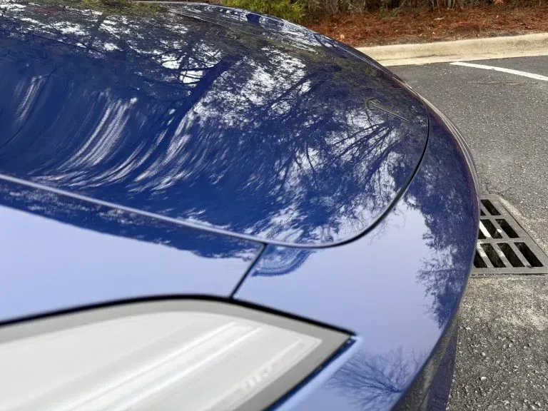 Blue car hood and headlight beside a street drain, with tree reflections on the glossy surface