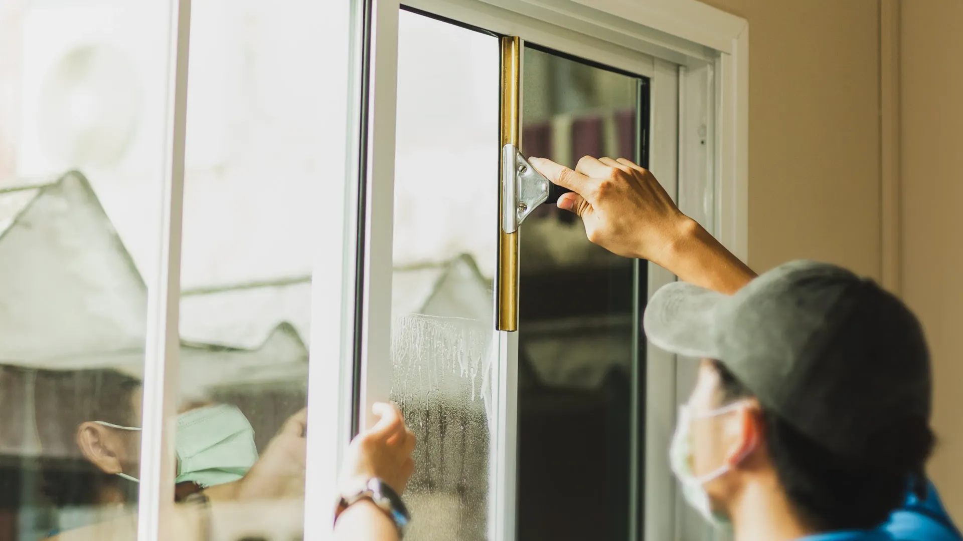 Person in a cap and mask cleaning a window with a cloth indoors.