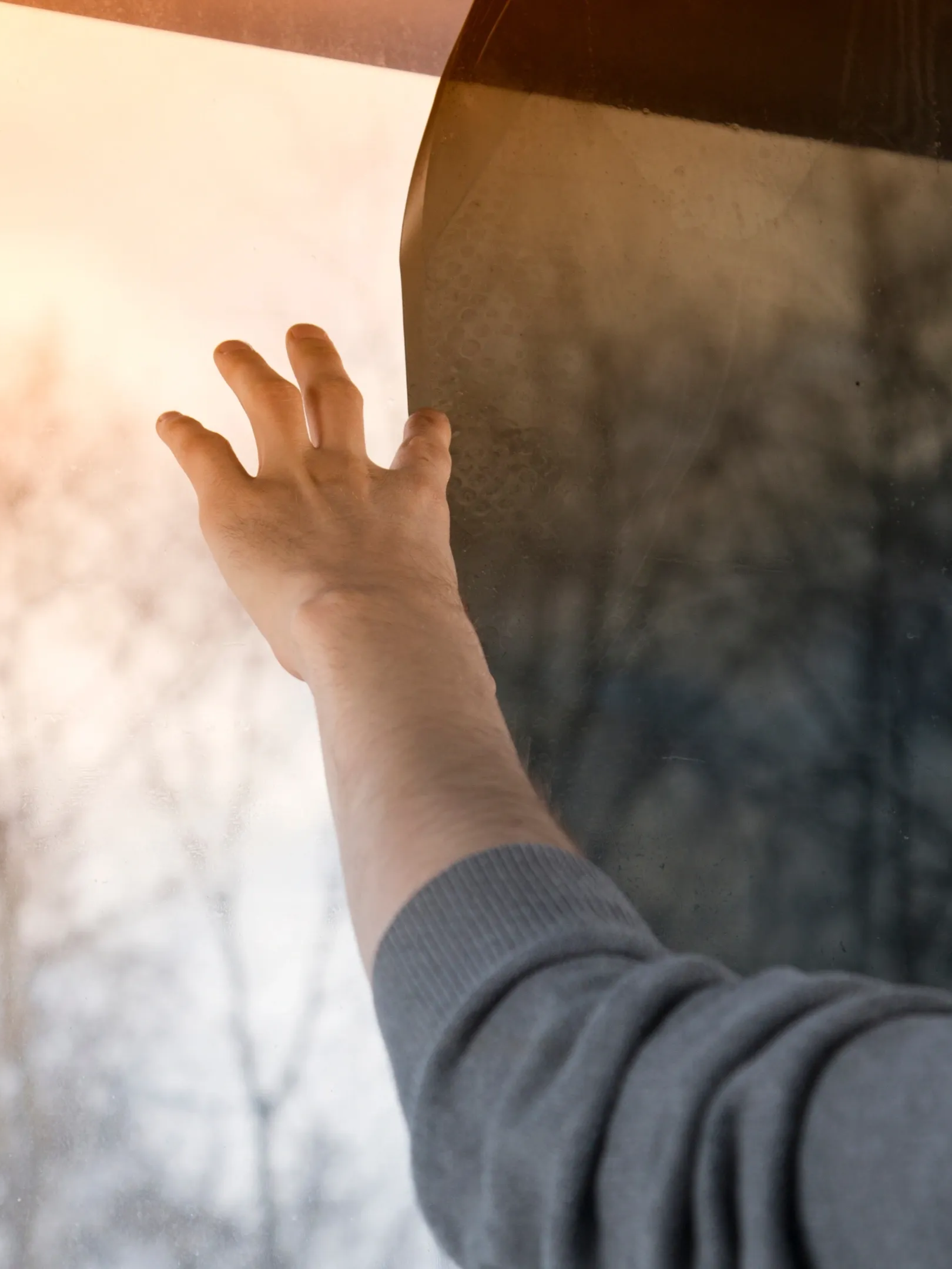 Hand reaching up toward bright sunlight through a window, with a gray sleeve visible