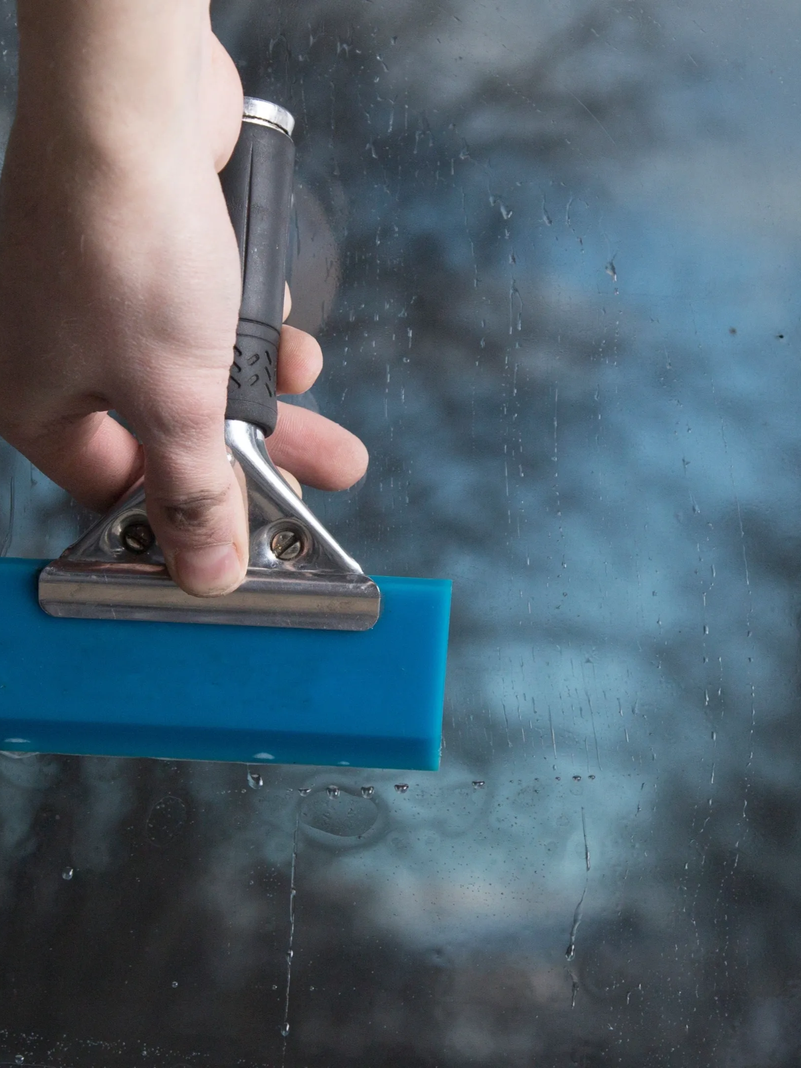 Hand pressing a squeegee across blue glass with water droplets on a window pane