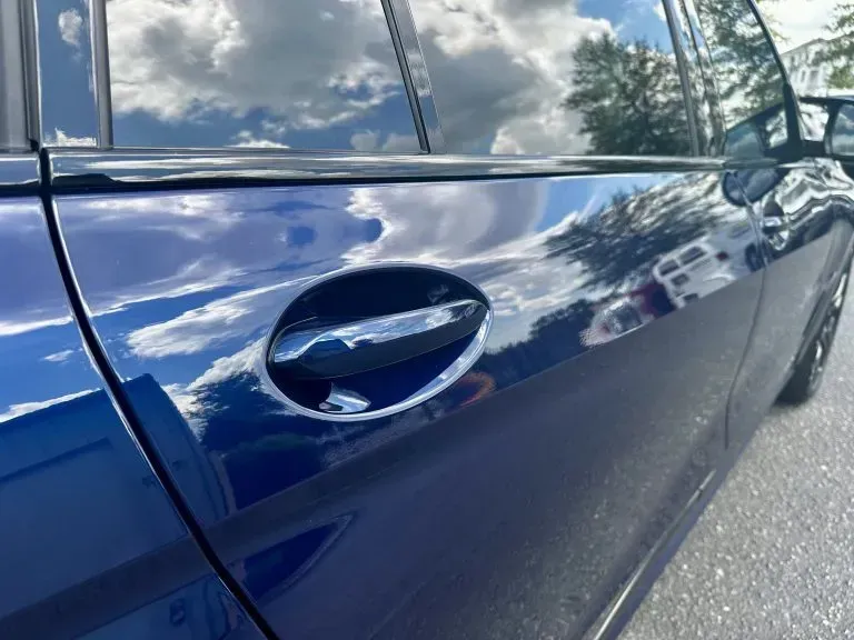 Close-up of a blue car door with a chrome handle reflecting clouds and trees
