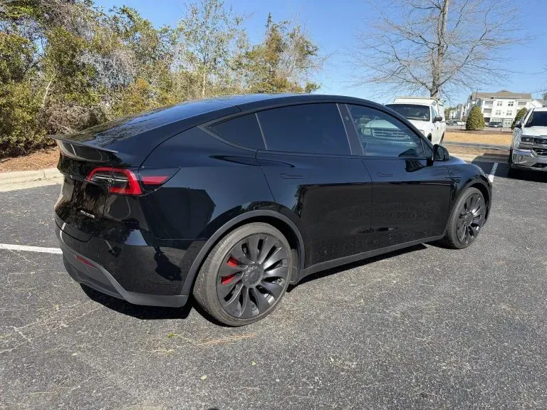 Black Tesla sedan parked on a lot, side view in daylight, with trees and other cars in the background