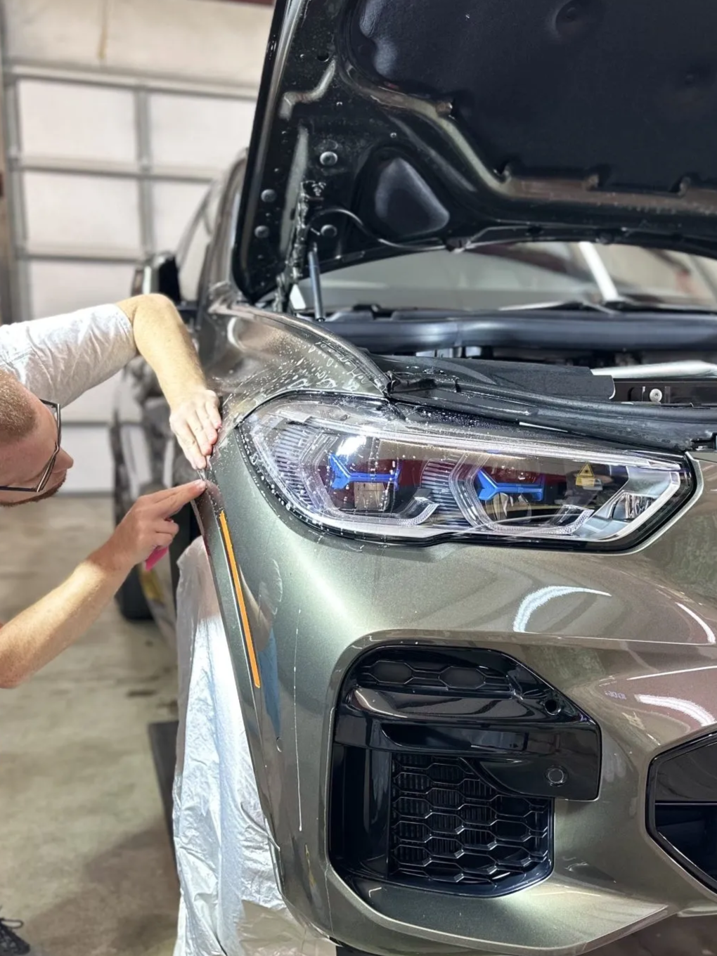 Person applying protective film to the front fender of a gray SUV in a garage