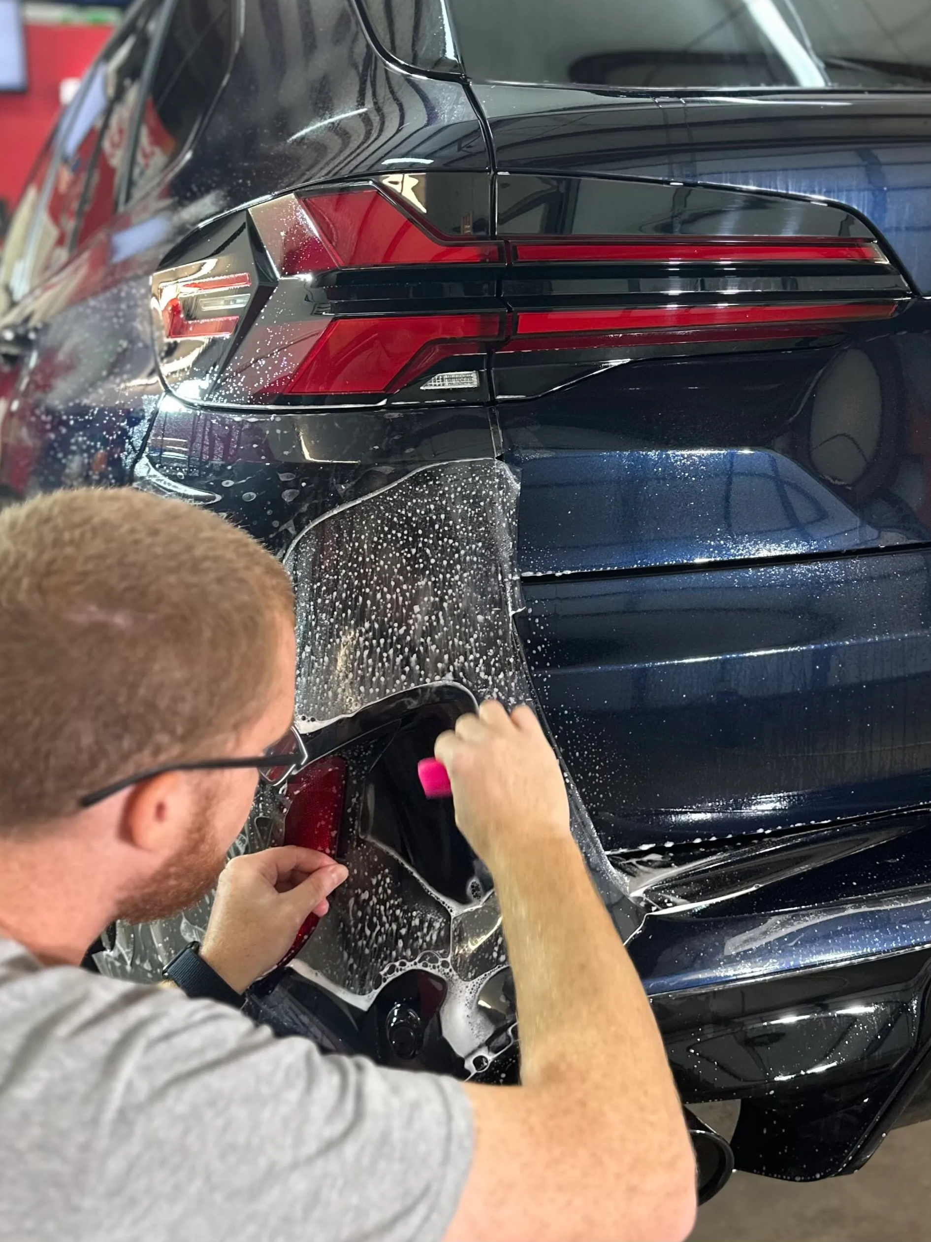 Person applying clear protective film to the rear of a dark blue car in a workshop