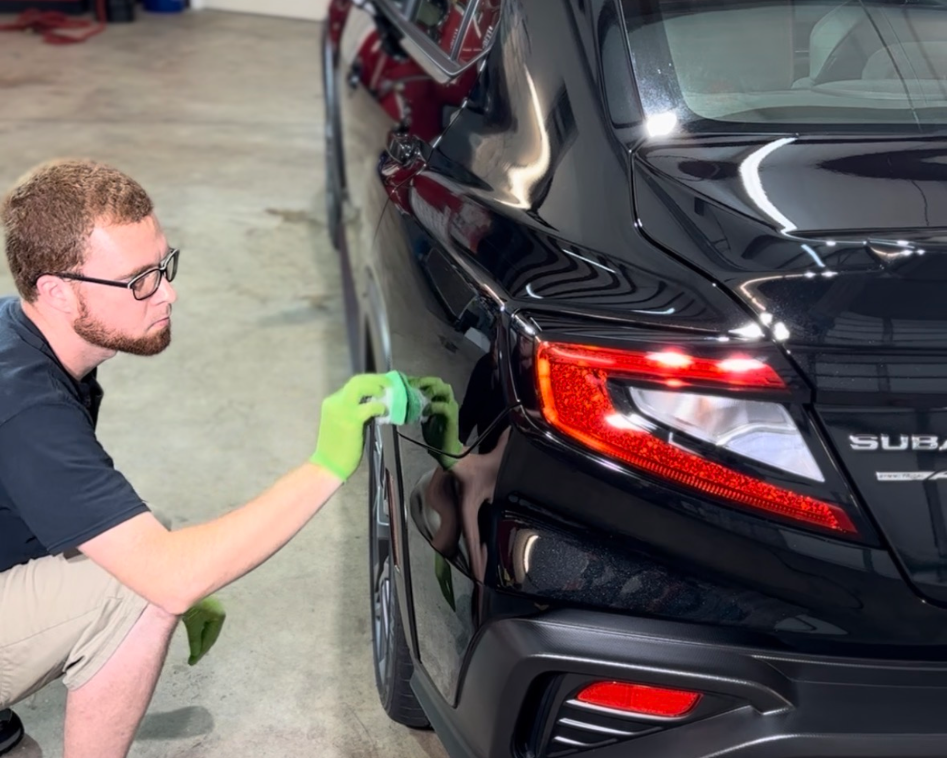 Worker polishing the rear of a black Subaru in a garage, wearing green gloves.