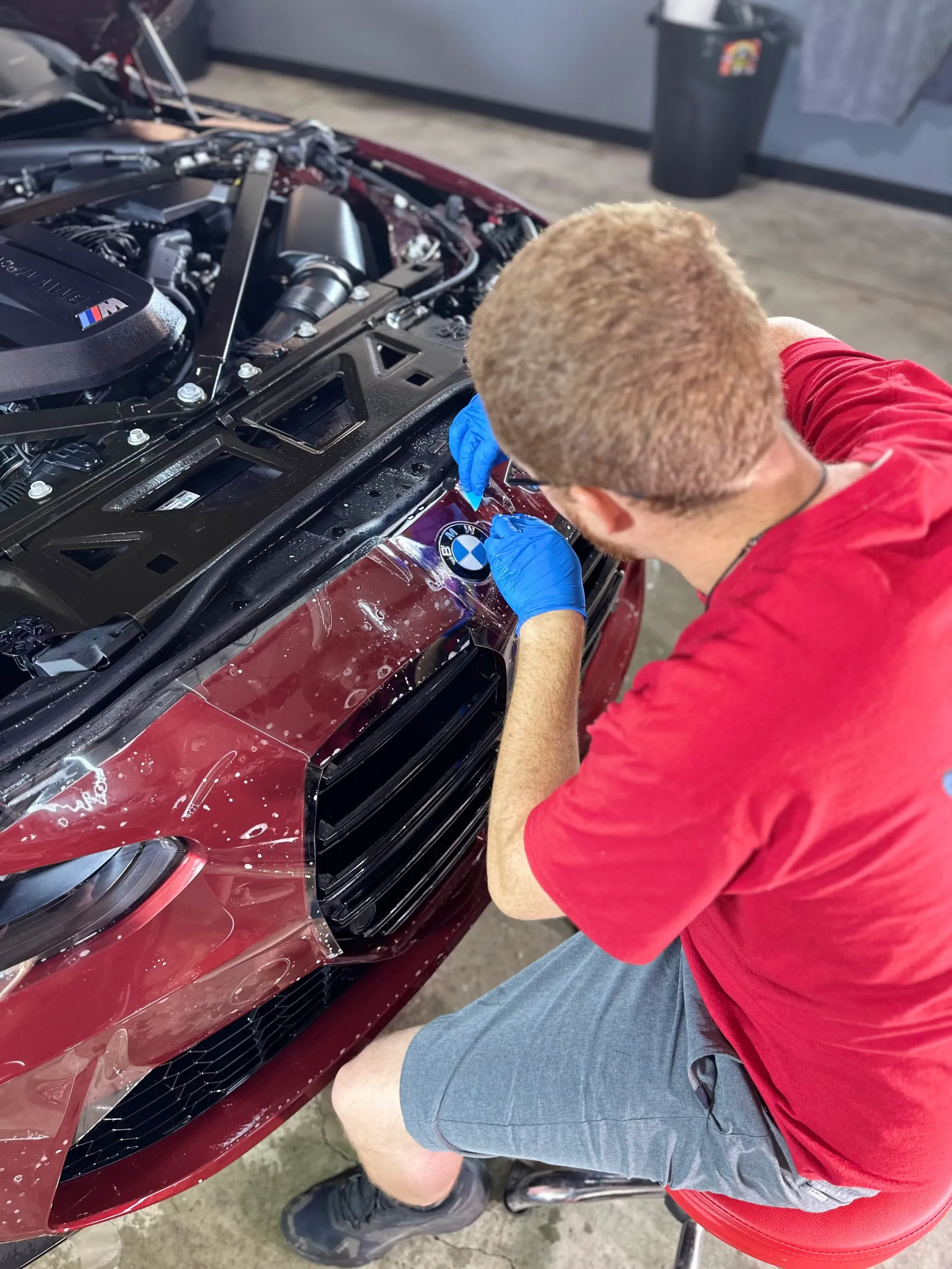 Person in a red shirt polishing a maroon car’s front bumper in a garage